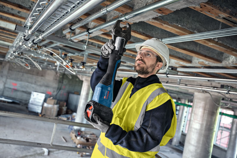 Eine Person mit Sicherheitsausrüstung verwendet ein Akku-Presswerkzeug zum Verbinden von Überkopfrohren. Anwendungsbild