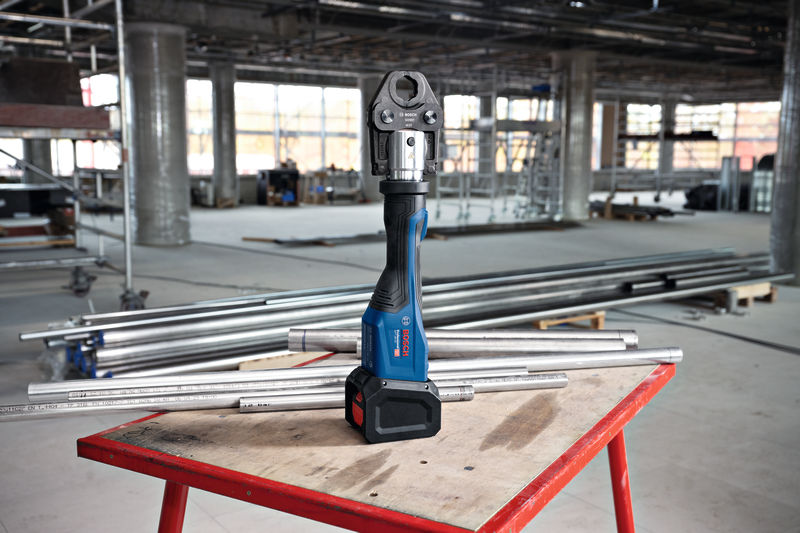 Ein Akku-Presswerkzeug steht auf einem Arbeitstisch, umgeben von Metallrohren in einer Baustelle. Anwendungsbild