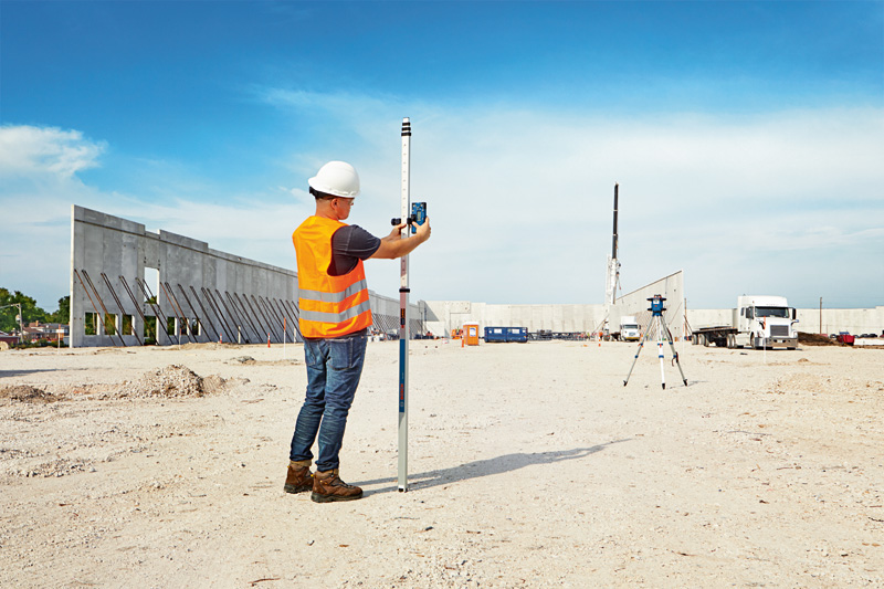 Eine Person mit Sicherheitsausrüstung verwendet auf einer Baustelle ein Lasernivelliergerät. Anwendungsbild