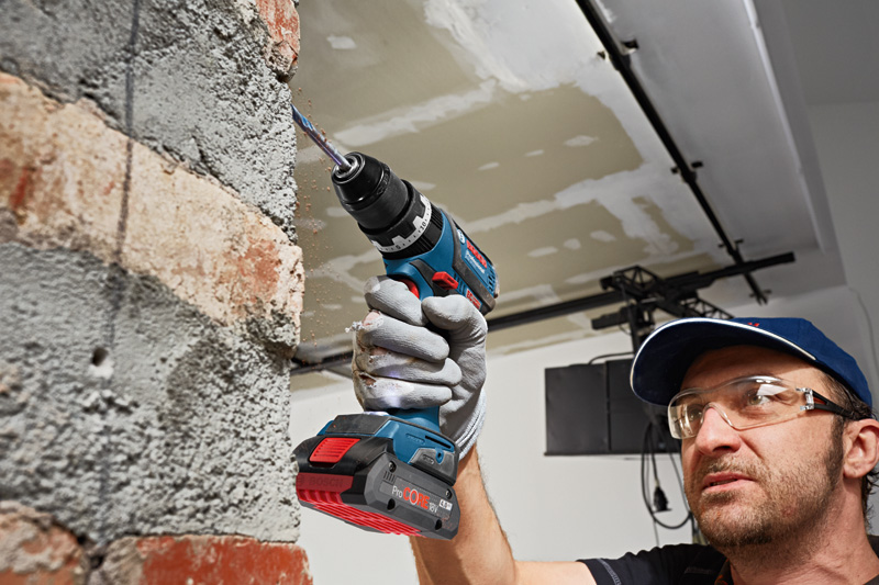 Eine Person mit Schutzausrüstung bohrt mit einer Akku-Bohrmaschine in eine Ziegelwand. Anwendungsbild
