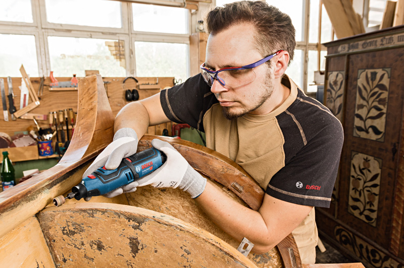 Eine Person mit Schutzausrüstung schleift in einer Werkstatt Holz mit einem kabellosen Rotationswerkzeug. Anwendungsbild