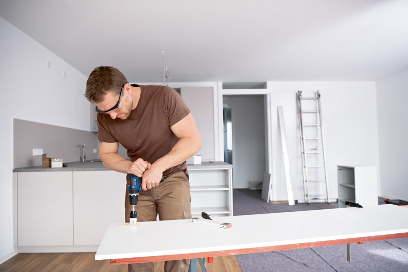 Eine Person mit Schutzausrüstung bohrt in einer modernen Küche in eine weiße Tafel. Anwendungsbild