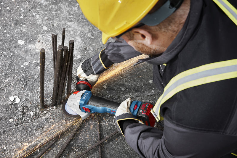 Eine Person mit Schutzausrüstung schleift mit einem Akku-Winkelschleifer Bewehrungsstahl auf Beton. Anwendungsbild