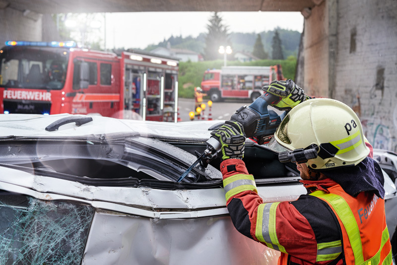 Eine Person mit Sicherheitsausrüstung schneidet mit einem Elektrowerkzeug ein beschädigtes Fahrzeug auf. Anwendungsbild