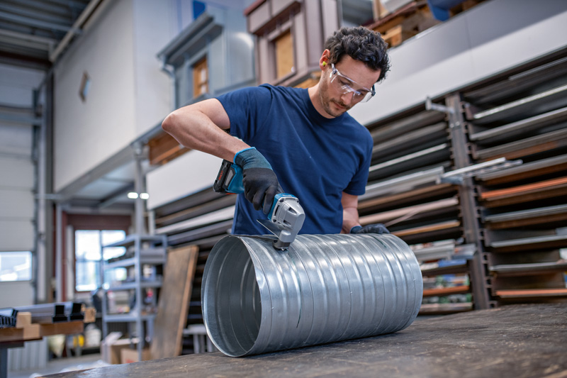 Eine Person mit Sicherheitsausrüstung schneidet mit einer kabellosen Metallschere einen großen Metallkanal durch. Anwendungsbild