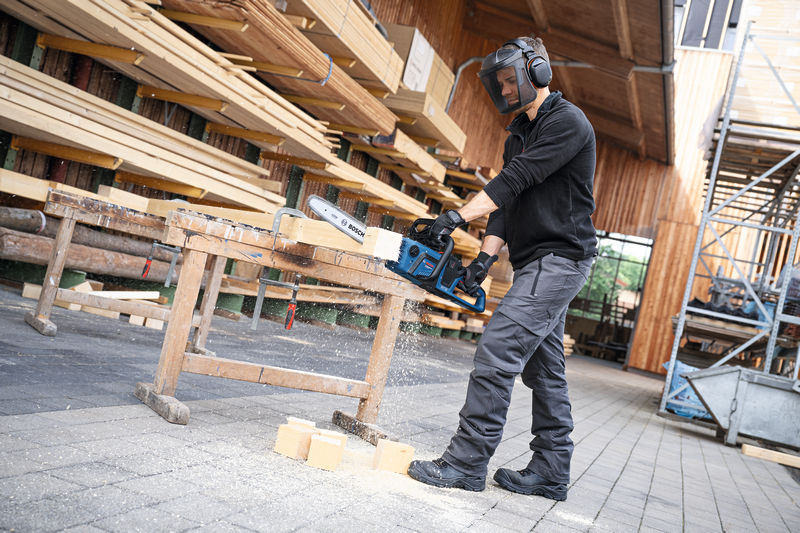 Eine Person mit Sicherheitsausrüstung sägt im Freien mit einer Akku-Kettensäge Holz. Anwendungsbild
