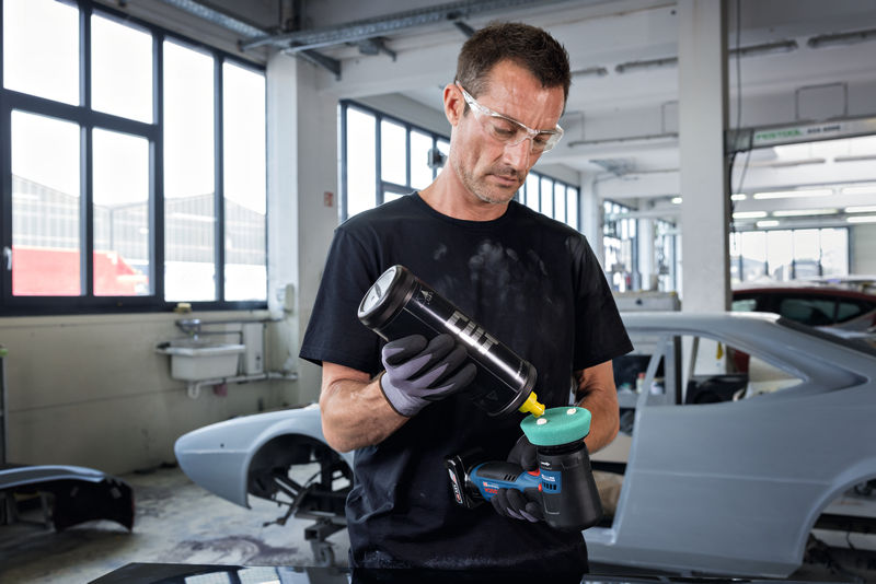 Eine Person mit Sicherheitsausrüstung trägt in einer Autowerkstatt Politur auf einer Poliermaschine auf. Anwendungsbild