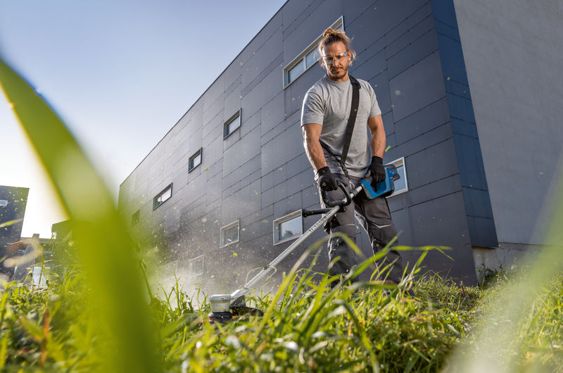 Eine Person mit Schutzausrüstung schneidet hohes Gras mit einem Akku-Rasentrimmer. Anwendungsbild