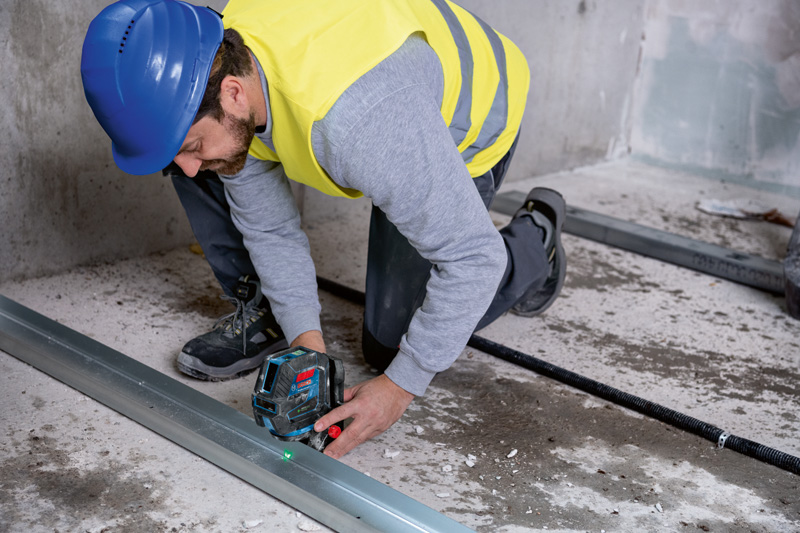 Eine Person mit Sicherheitsausrüstung richtet einen Metallrahmen mit einem Lasernivelliergerät auf einem Betonboden aus. Anwendungsbild