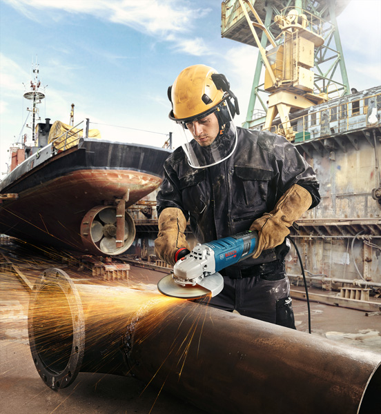 Eine Person mit Sicherheitsausrüstung schleift auf einer Werft ein großes Metallrohr. Anwendungsbild Weitwinkelaufnahme