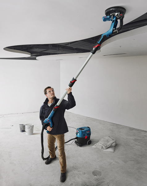 Eine Person mit Schutzausrüstung schleift in einem Renovierungsraum eine Decke mit einem Trockenbauschleifer. Anwendungsbild