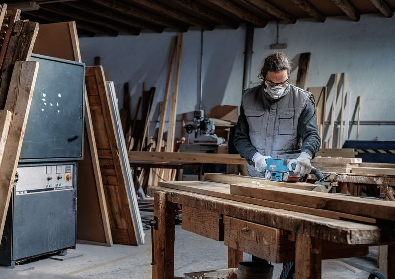 Eine Person mit Schutzausrüstung schleift in einer Werkstatt mit einem Elektrowerkzeug ein Holzbrett. Anwendungsbild