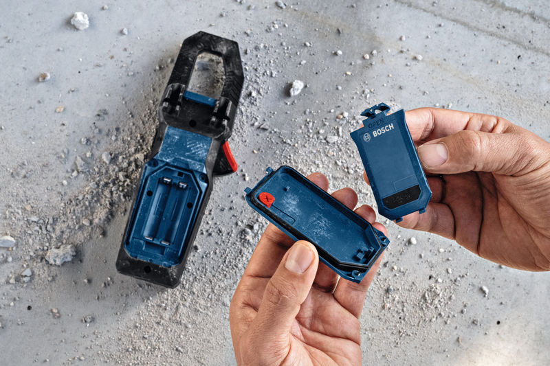 Person öffnet ein blaues Batteriefach neben einem Werkzeug auf einem staubigen Betonboden. Anwendungsbild
