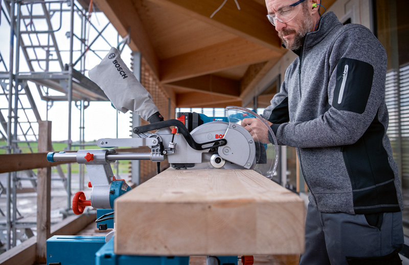 Eine Person mit Sicherheitsausrüstung bedient eine kabellose Gehrungssäge, um einen Holzbalken zu schneiden. Anwendungsbild