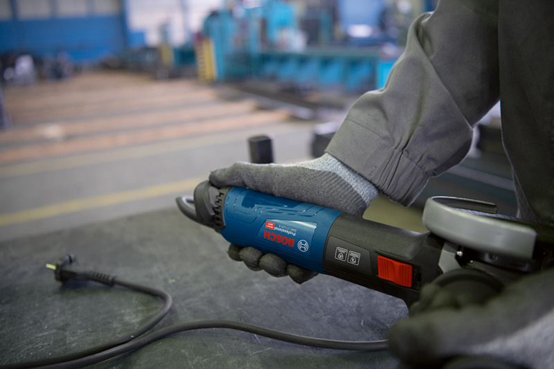 Eine Person mit Schutzausrüstung hält einen Winkelschleifer auf einer Metallwerkbank. Anwendungsbild