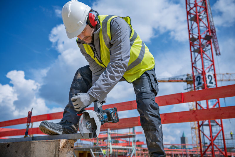 Eine Person mit Schutzausrüstung schneidet auf einer Baustelle Metall mit einem Akku-Winkelschleifer. Anwendungsbild
