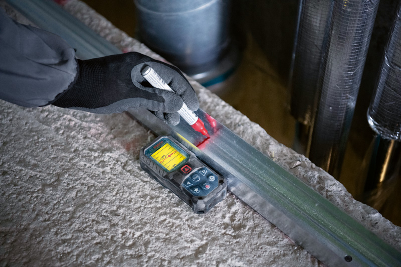 Eine Person mit Sicherheitsausrüstung markiert einen Metallträger mit einem Lasermessgerät. Anwendungsbild